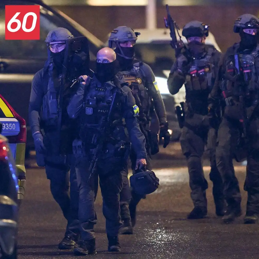 A group of heavily armed, masked police officers in tactical gear walk at night, one in the center looks directly at the…