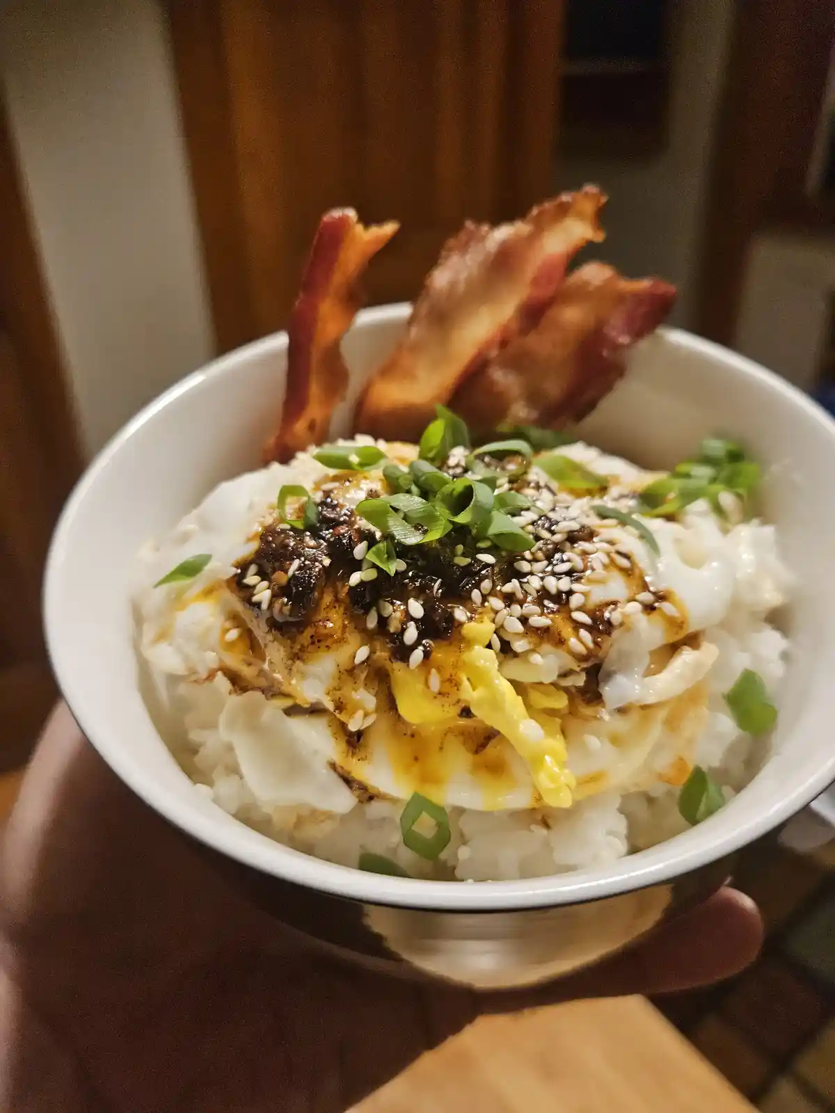 A close-up photo shows a white bowl held in a hand, containing a rice bowl topped with a fried egg, drizzled with dark…