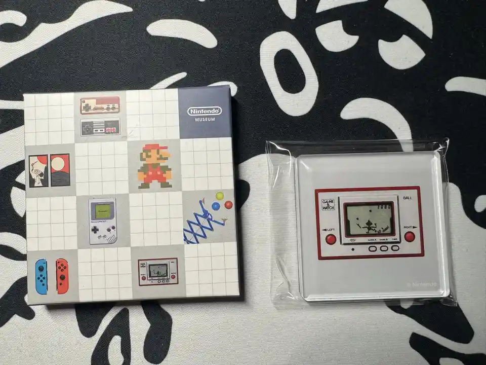A silver Nintendo Game & Watch handheld game in its clear plastic packaging sits next to a Nintendo Museum collectible box…