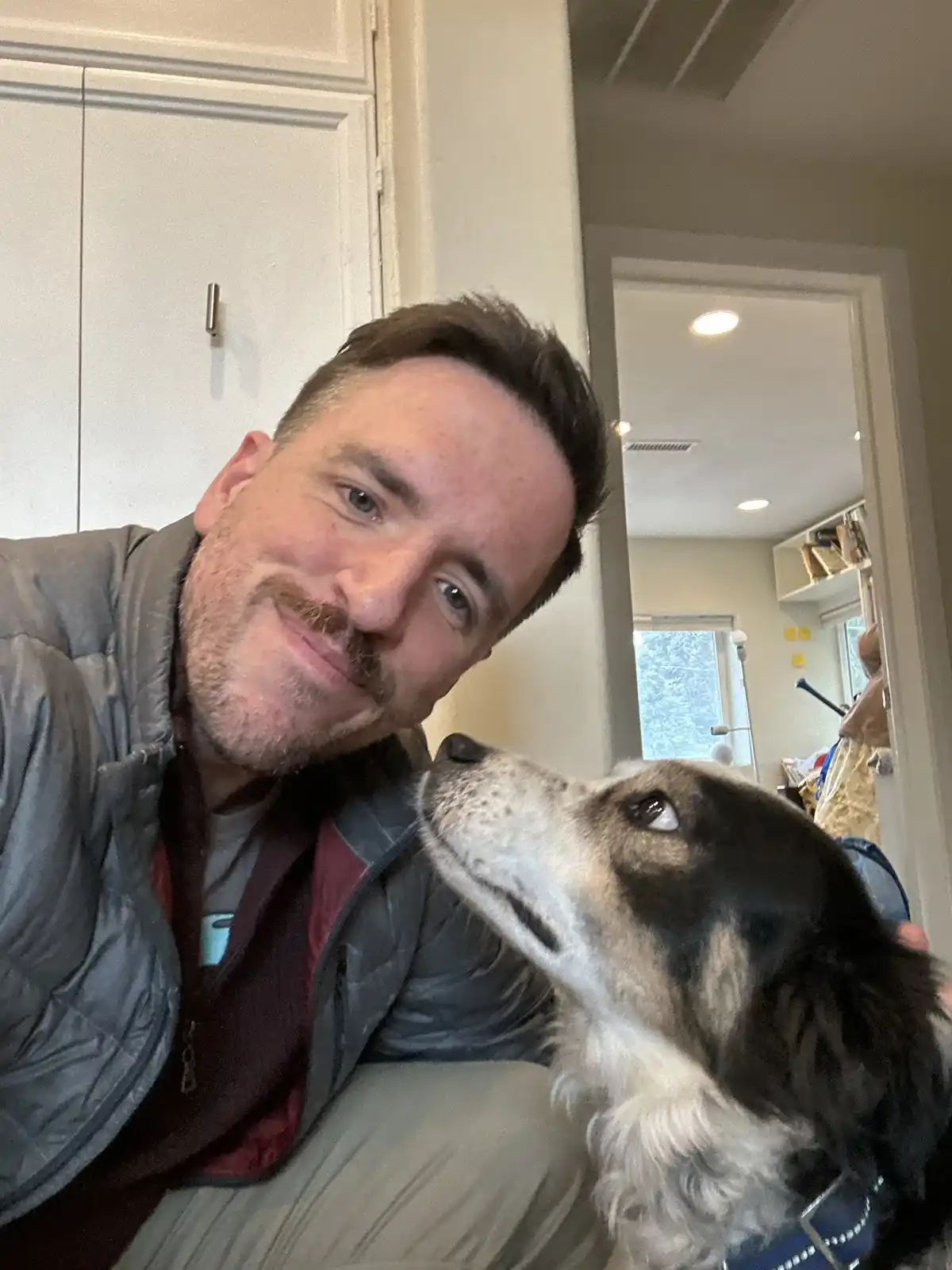 A man with a mustache and gray jacket smiles at the camera while holding a black and white dog that looks up at him. The dog…