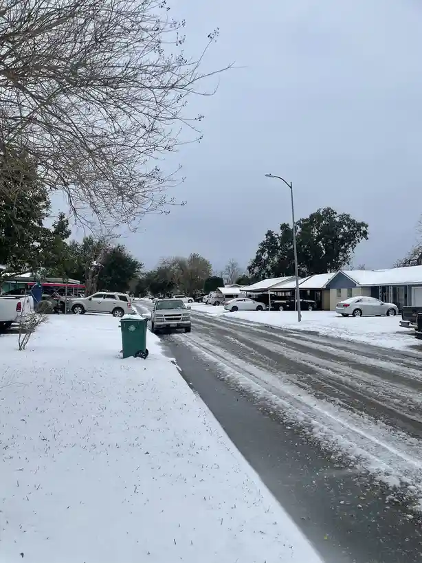 A residential street in a suburban neighborhood is covered in snow, with cars parked along the sides and a green trash can…