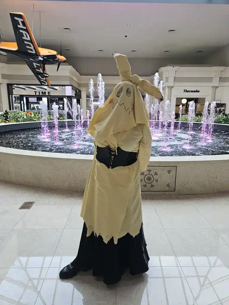 A person in a yellow, tattered ghost costume with a black corset and black skirt stands in a mall atrium facing a fountain…
