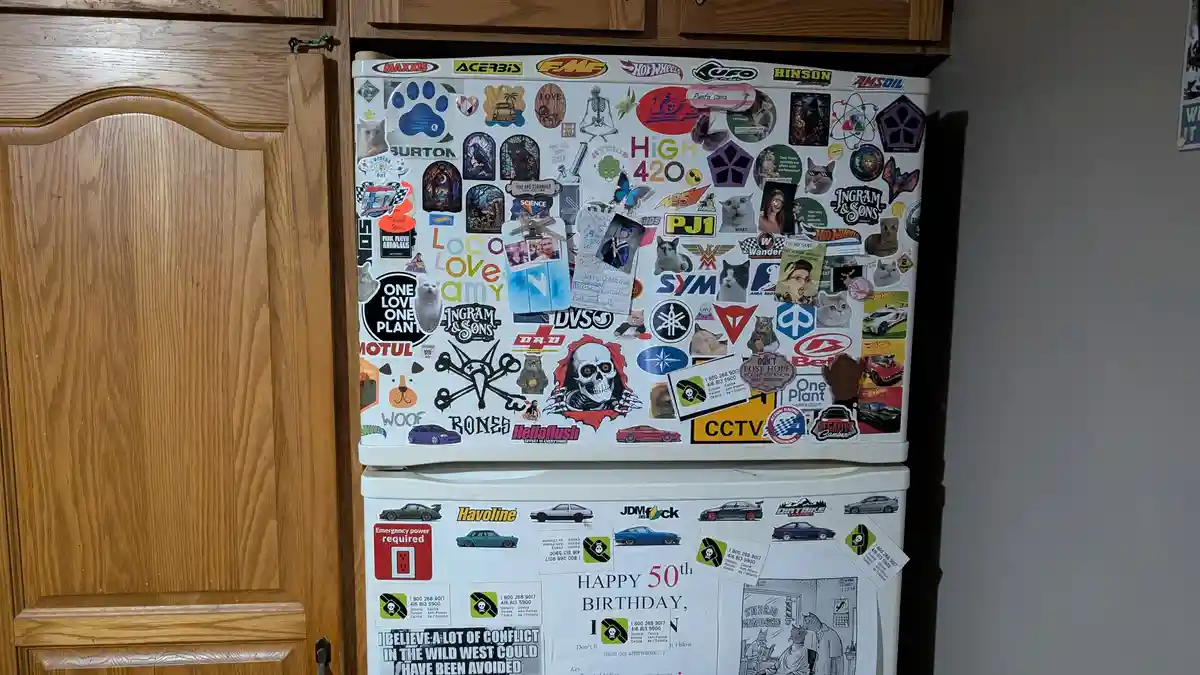 A white refrigerator is covered in a dense collage of stickers, including car brands, cannabis-related imagery, cats…