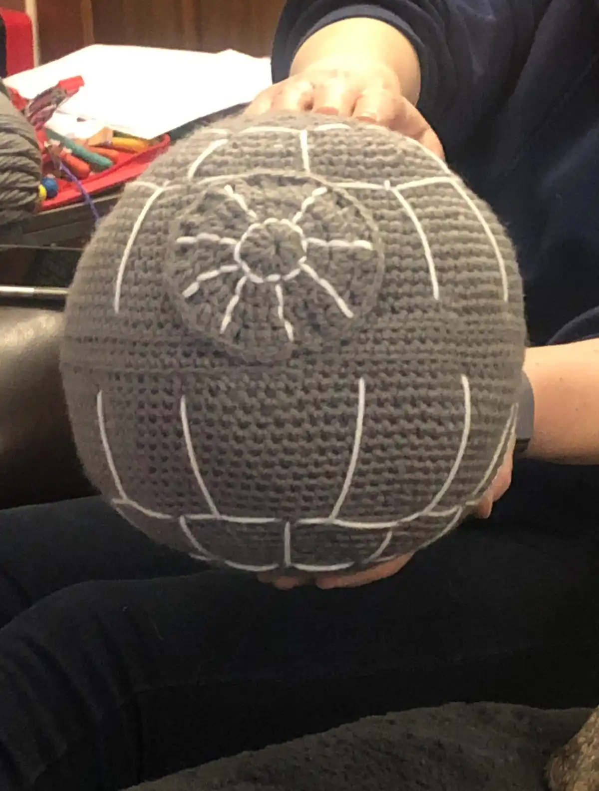 A person holds a gray, crocheted spherical object resembling the Death Star from Star Wars, featuring white stitched lines…