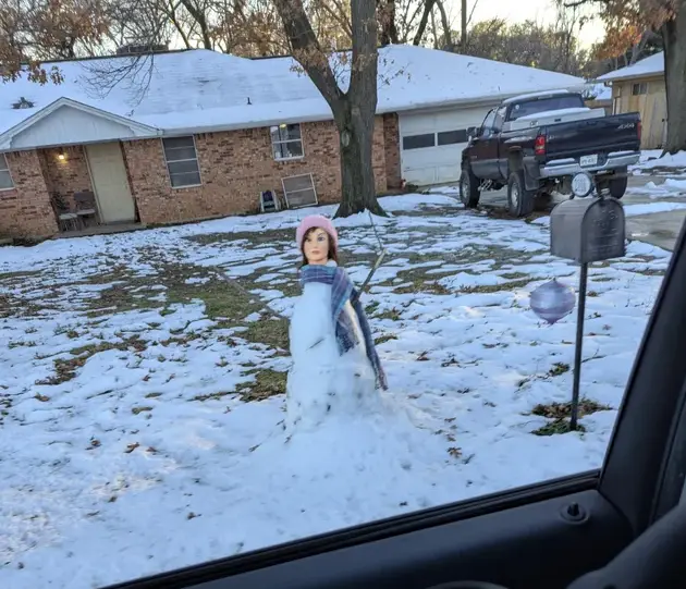 A photo taken from inside a car shows a snowman in a snowy yard, constructed from the head of a mannequin wearing a pink hat…