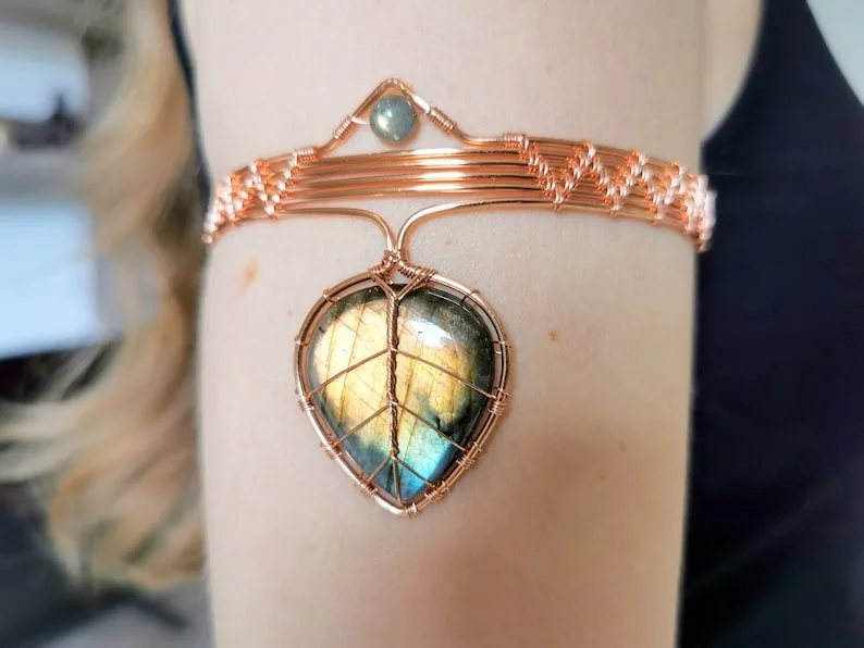 A close-up photo shows a person's forearm wearing a copper wire-wrapped bracelet with a large, teardrop-shaped labradorite…