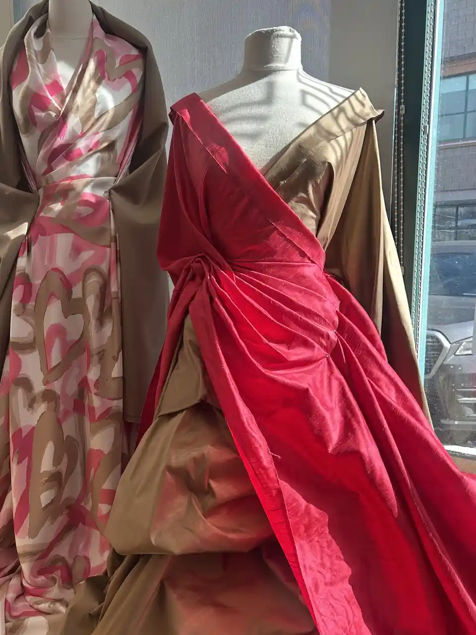 Two mannequins display draped, avant-garde dresses—one with pink and beige abstract patterns, the other with a bold red and…