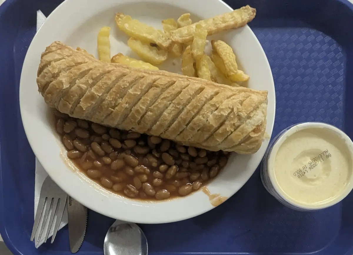 A plate of food on a blue tray includes a large pastry roll, baked beans, and french fries, with a small container of sauce…