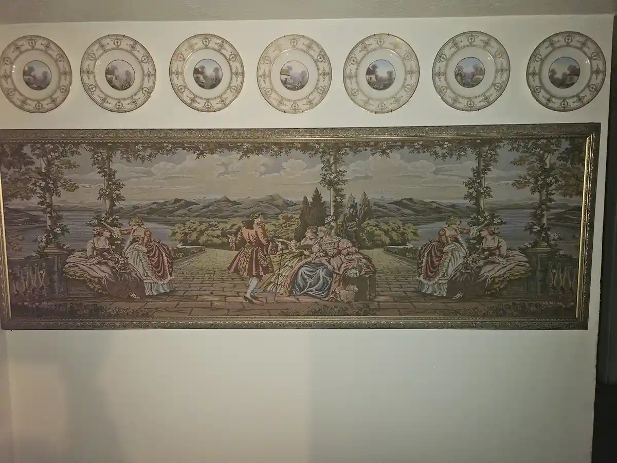 A wall displays a large, ornate framed tapestry depicting aristocrats in 18th-century attire on a terrace overlooking water…