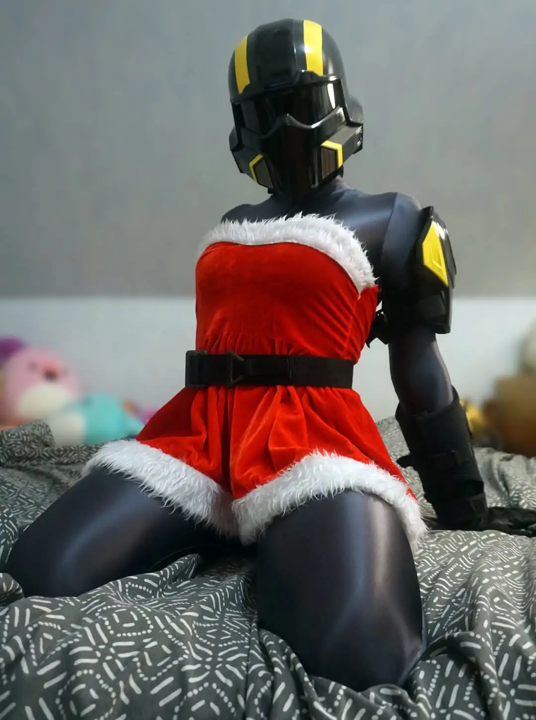 A person in a black and yellow armored helmet and suit is sitting on a bed wearing a red velvet Santa dress with white fur…
