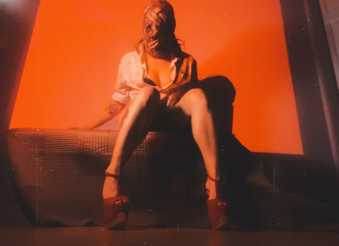 A person with a bandaged, obscured face sits on a textured couch under intense orange lighting, wearing a white shirt, dark…