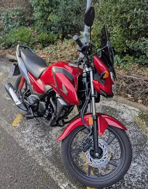 A red Honda motorcycle is parked on a paved surface next to green bushes, with visible details like the front wheel…