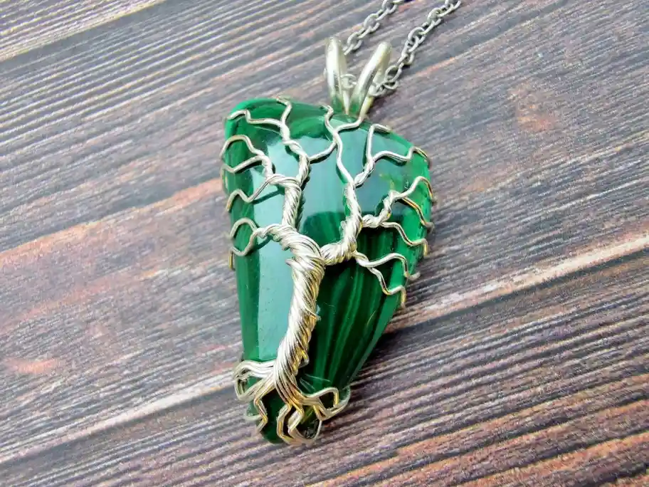 A green, heart-shaped malachite pendant with a silver wire tree of life design is displayed on a dark wooden surface. The…