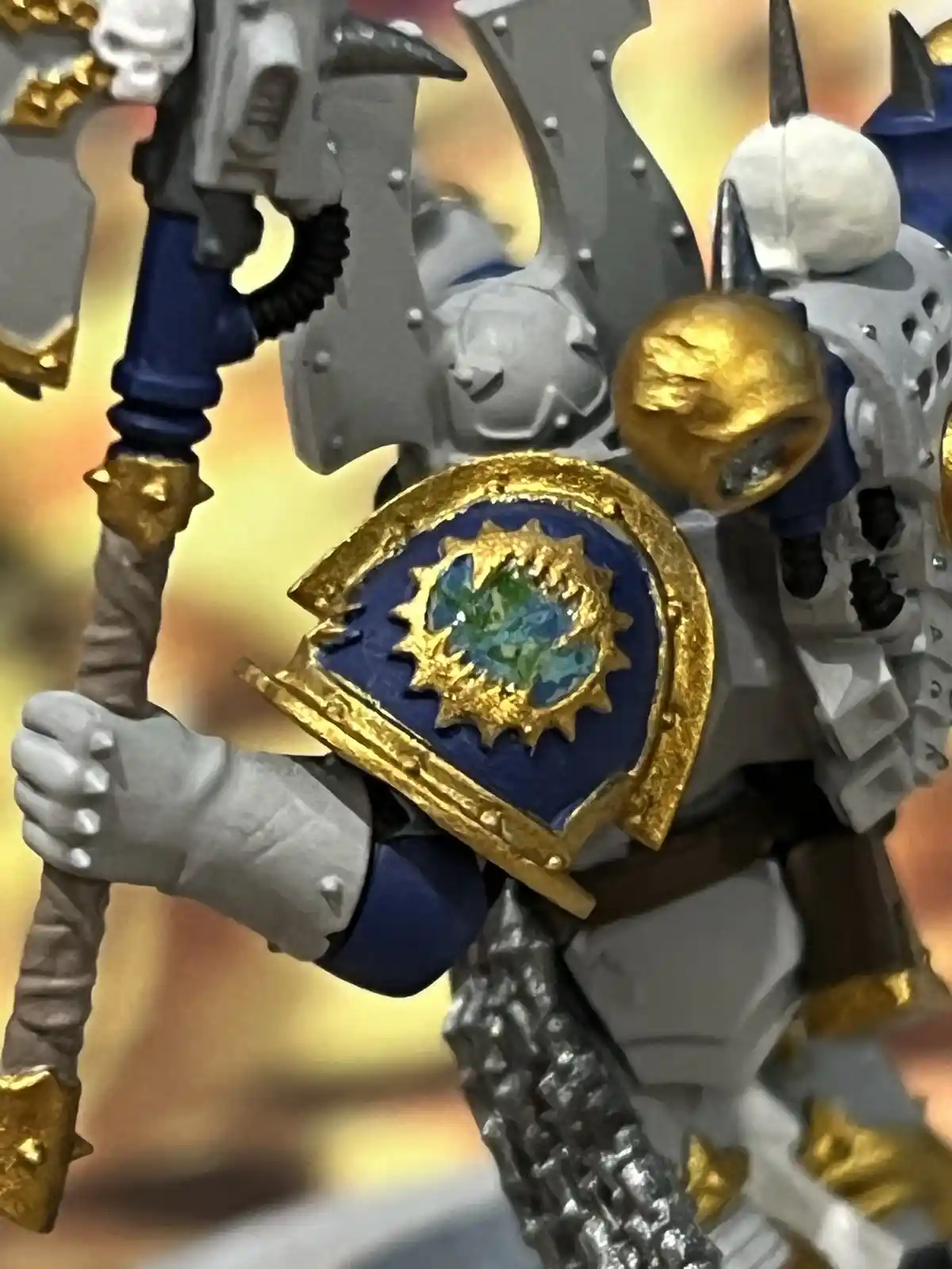 A close-up of a painted miniature warrior figure, likely from Warhammer 40,000, showing a shield with a blue field and gold…