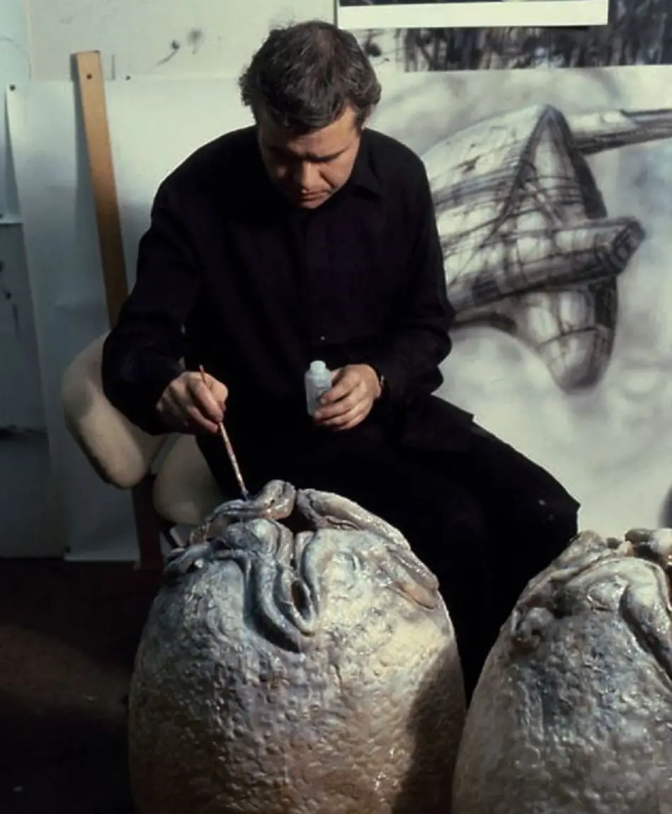 A man in a black shirt sits in an art studio, intently painting a large, textured sculpture of an octopus with a brush…