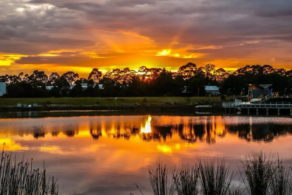 A serene sunset over a calm body of water, with vibrant orange and yellow hues reflecting in the surface. Silhouettes of…