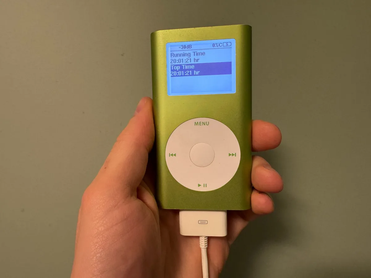 A hand holds a green first-generation iPod with a white USB cable plugged into its bottom. The screen displays 'Running Time…