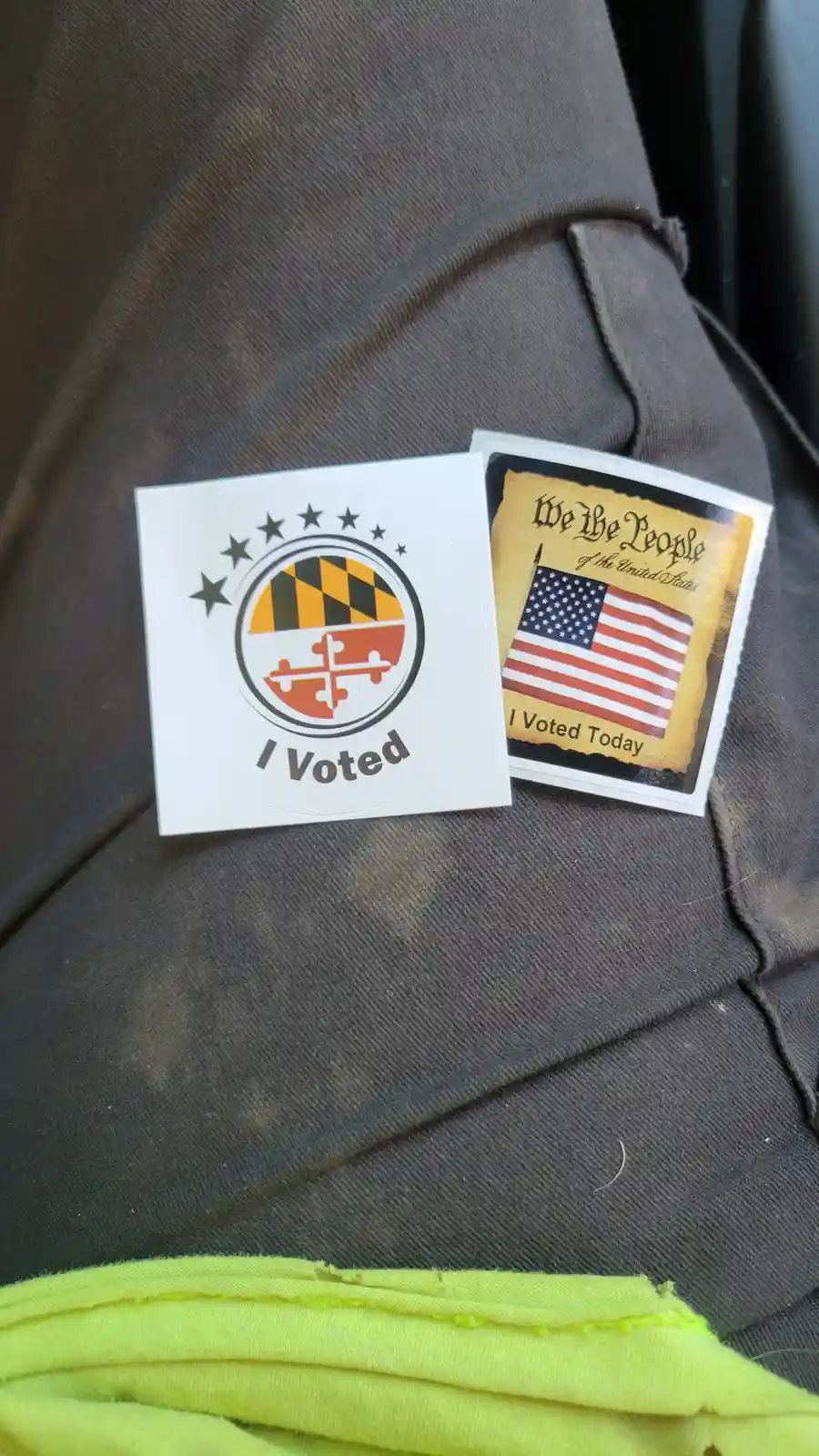 Two 'I Voted' stickers rest on a person's dark gray pants. One features the Maryland state flag and says 'I Voted'. The…