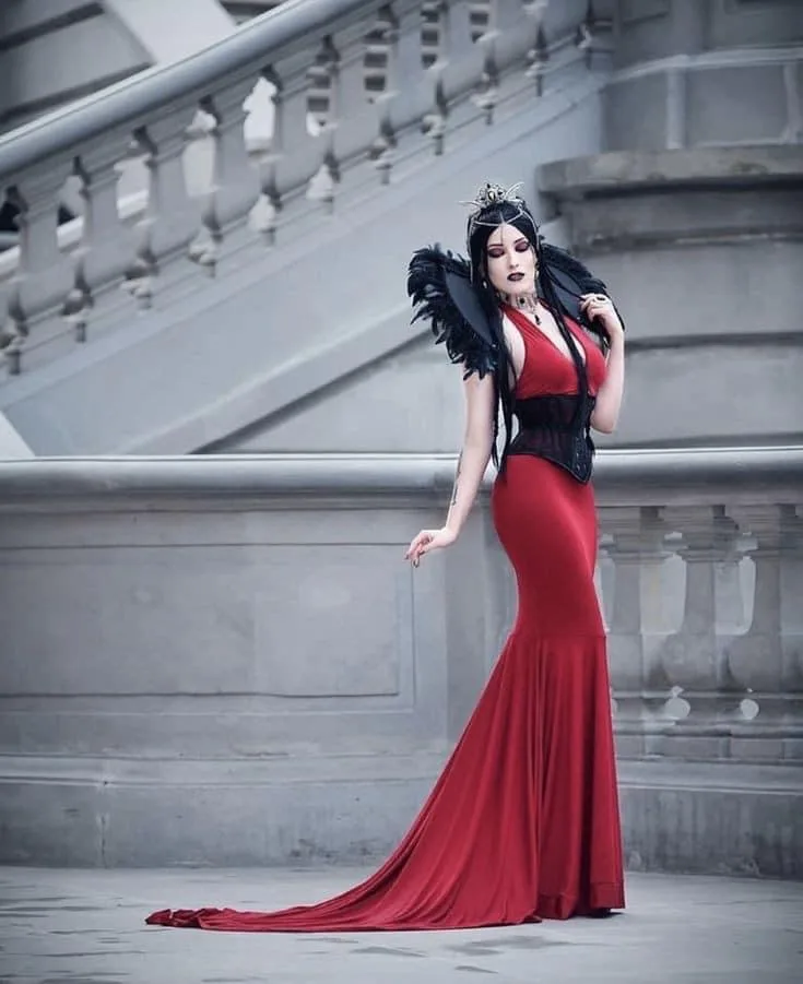 A woman in a striking red mermaid-style gown with black lace detailing and dramatic black feathered shoulder pieces poses on…