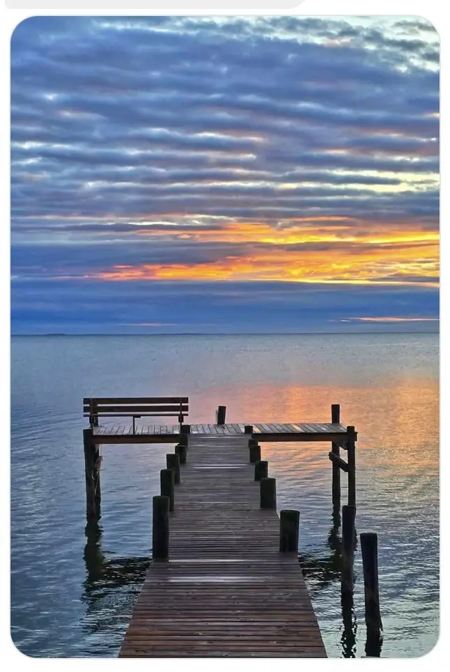 A serene photograph of a wooden pier extending into calm water under a dramatic sunset sky with orange and blue hues. A…