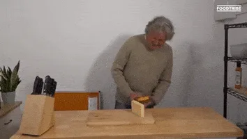 A man in a beige sweater stands at a wooden table with two blocks of cheese, one yellow and one white, arranged vertically…