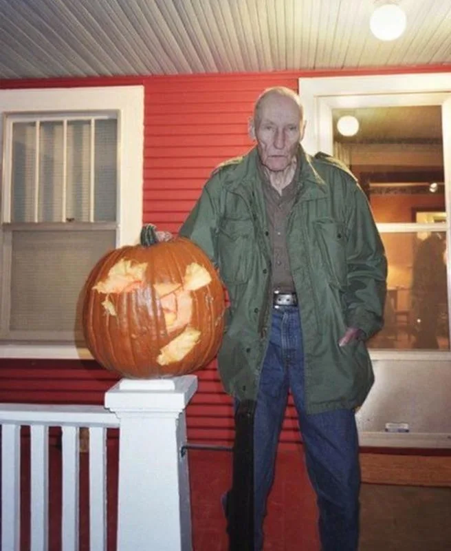 An elderly man in a green jacket and jeans stands on a porch next to a large, carved pumpkin that has been cut open and…