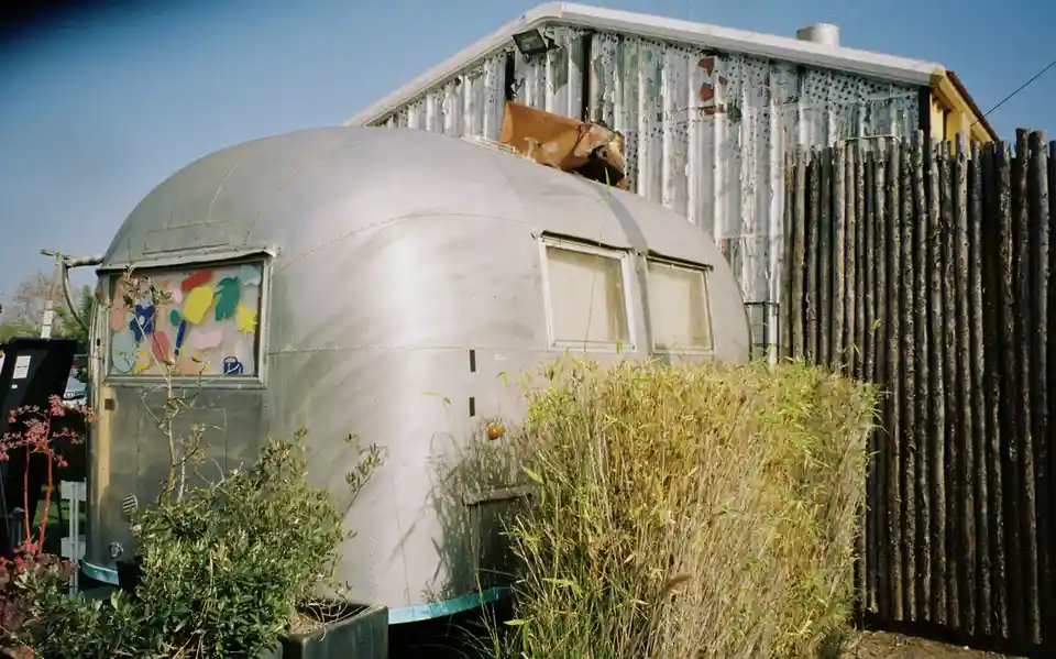A vintage silver Airstream trailer sits in a yard, partially obscured by overgrown bushes and plants, with a weathered…