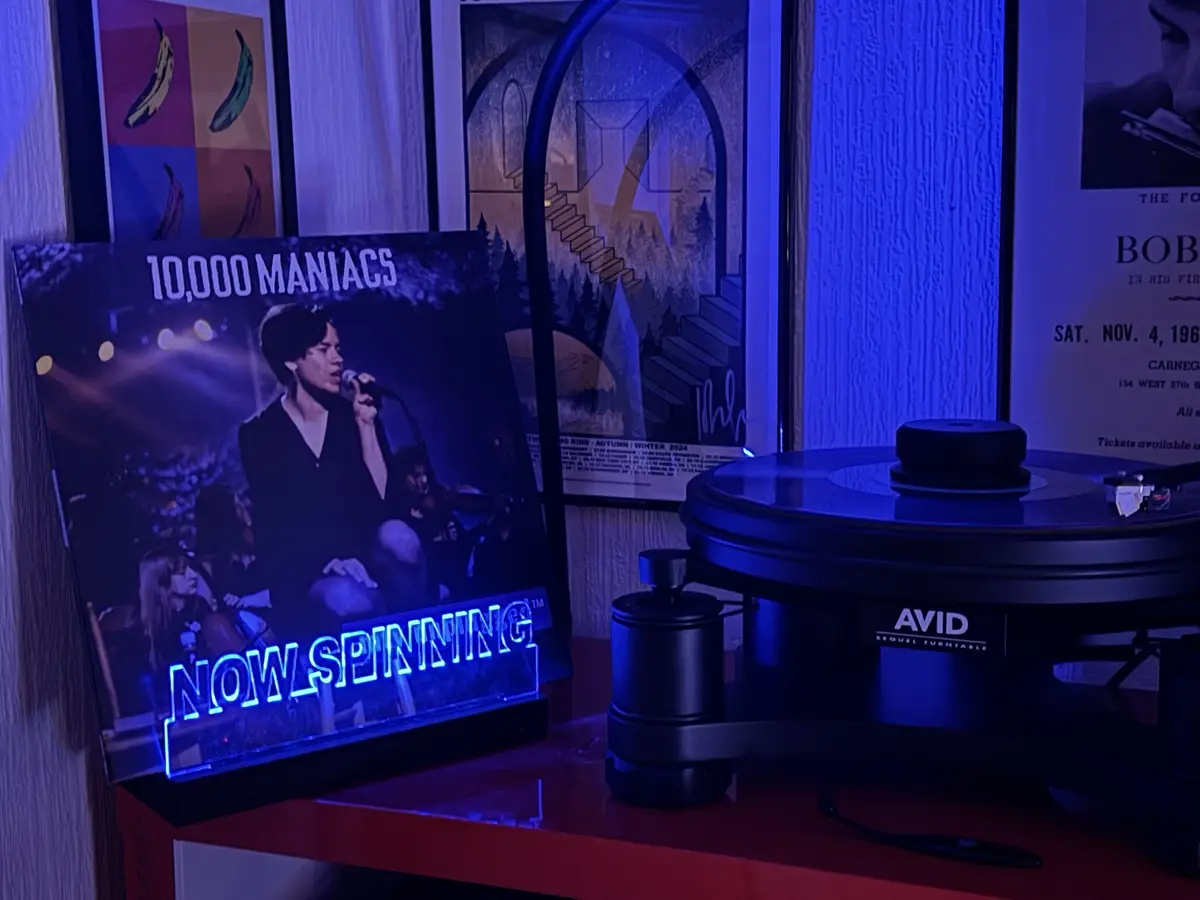 A vinyl record player sits on a red shelf next to a glowing neon sign that reads 'NOW SPINNING' over a poster for the band…