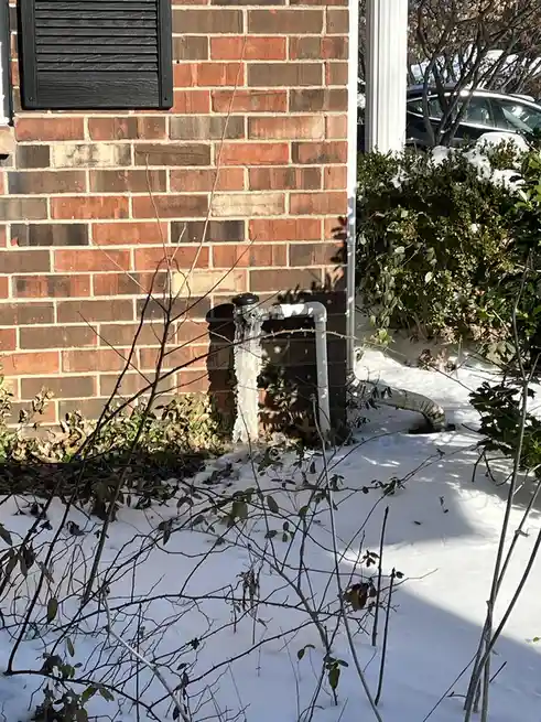 An outdoor photo shows a white PVC pipe protruding from a brick wall, with a large icicle hanging from its top, surrounded…