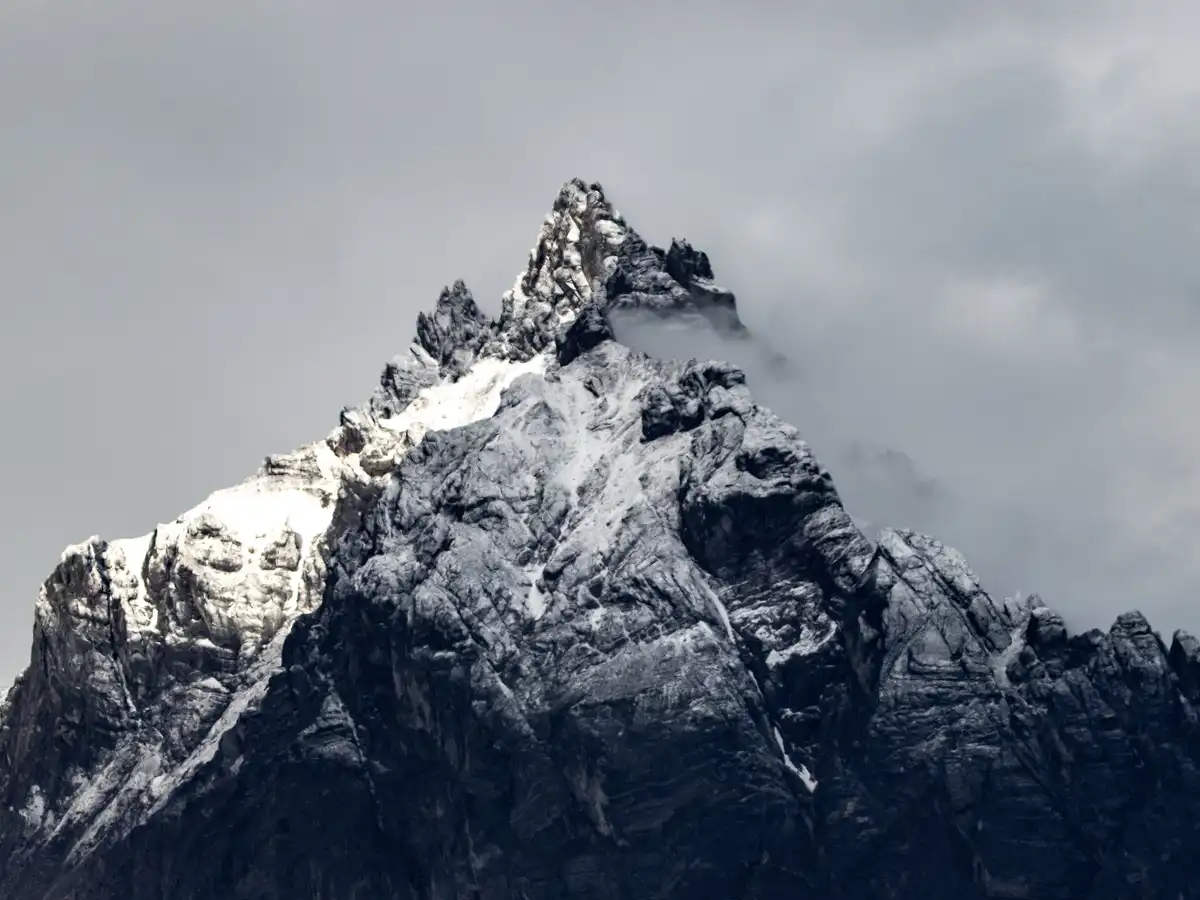 A dramatic, snow-dusted mountain peak rises sharply against a cloudy, overcast sky, its jagged ridges and rocky textures…