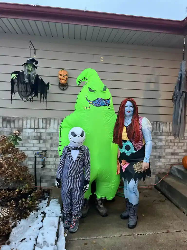 Three people dressed as Halloween characters stand on a snowy porch: a child in a Jack Skellington costume, a giant…