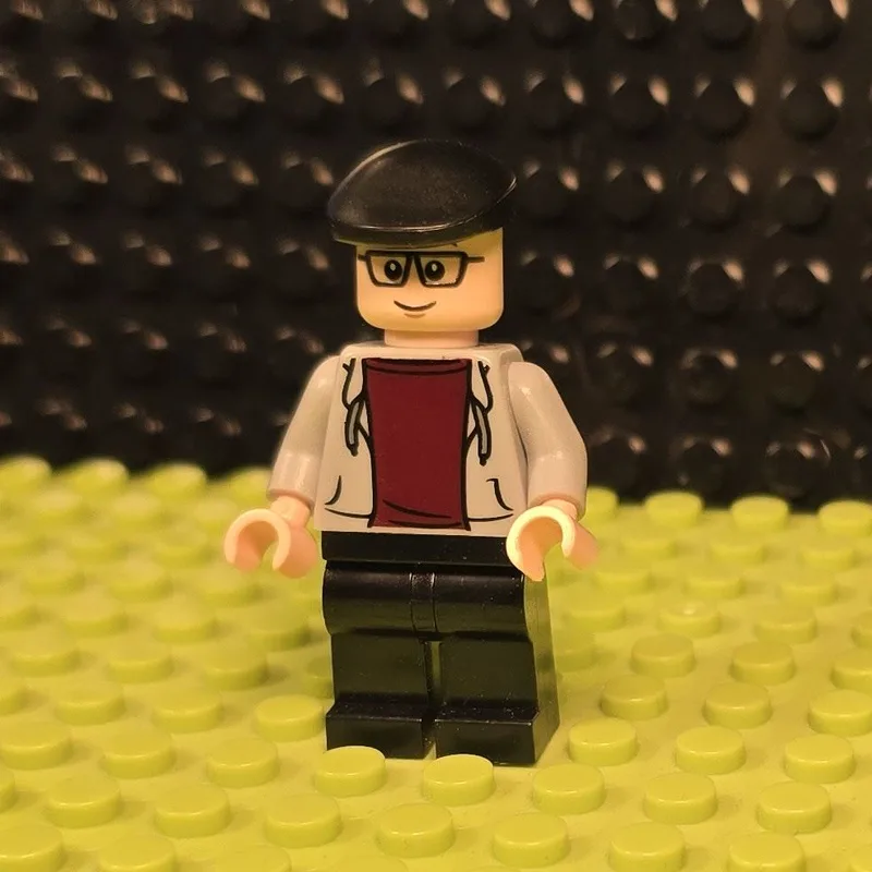 A lego minifigure wearing a black beret, glasses, a white jacket over a red shirt, and black pants stands on a lime green…