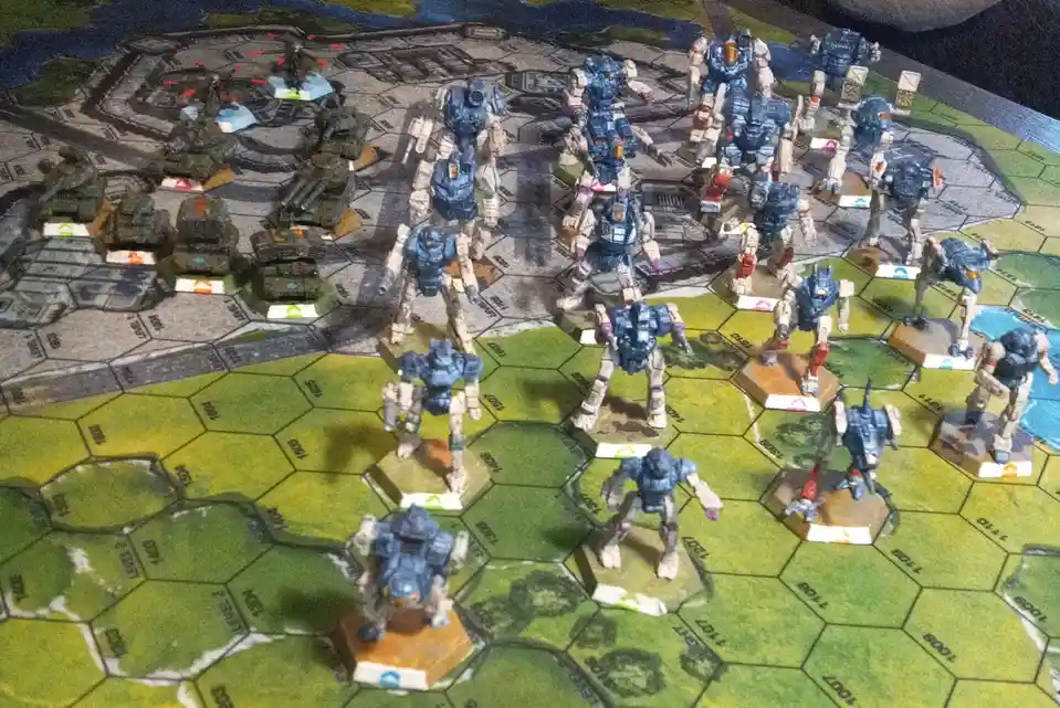 A tabletop wargame setup shows numerous blue and grey mecha and tank miniatures arranged on a hexagonal grid map. The text…