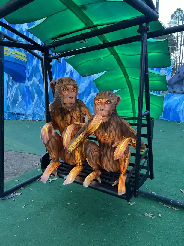 Two illuminated monkey lantern sculptures sit side-by-side on a black metal bench under a green canopy, one holding a banana…