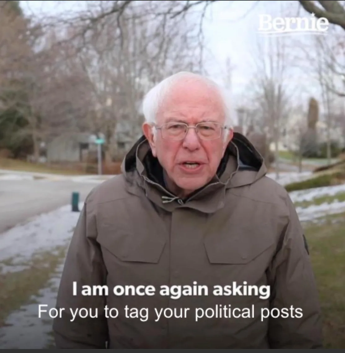 An older man with white hair and glasses, wearing a brown hooded jacket, stands outdoors in a snowy, tree-lined area. The…