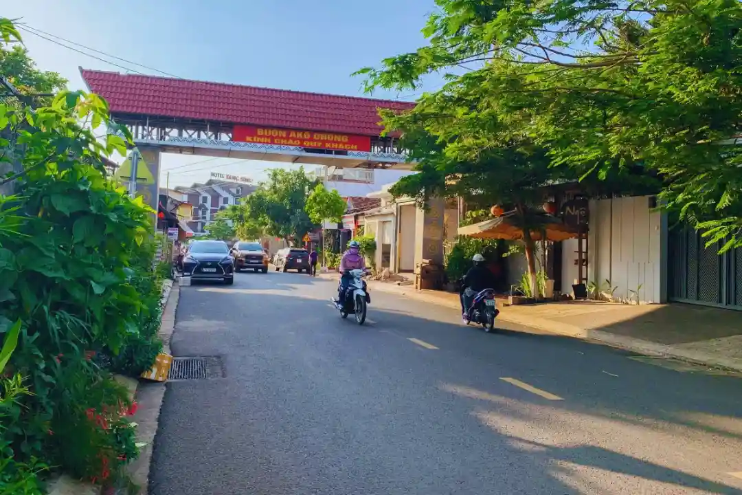 A street scene in Buon Ako Dong, Vietnam, featuring a red-roofed archway with yellow text reading 'BUON AKO DRONG KINH CHAO…