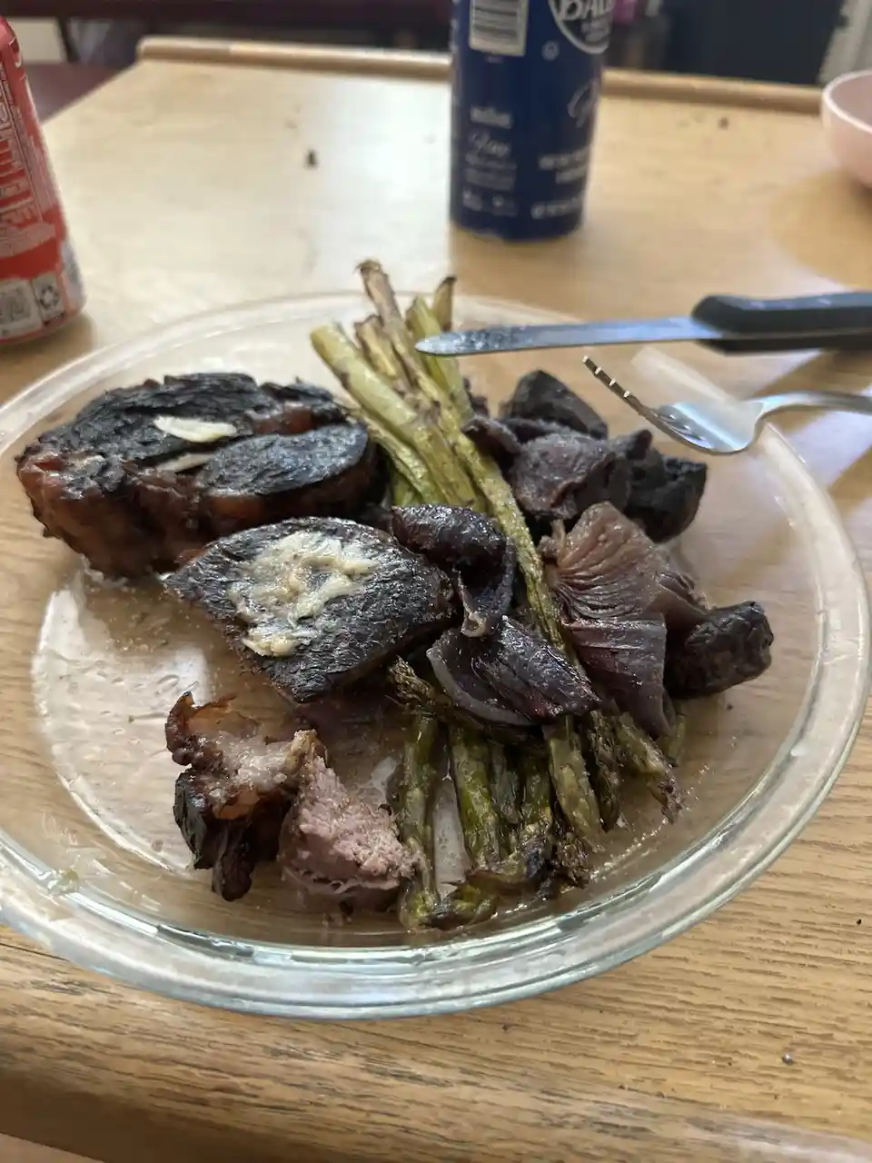 A close-up photo of a clear glass plate on a wooden table holding a meal that includes charred, blackened meat slices…