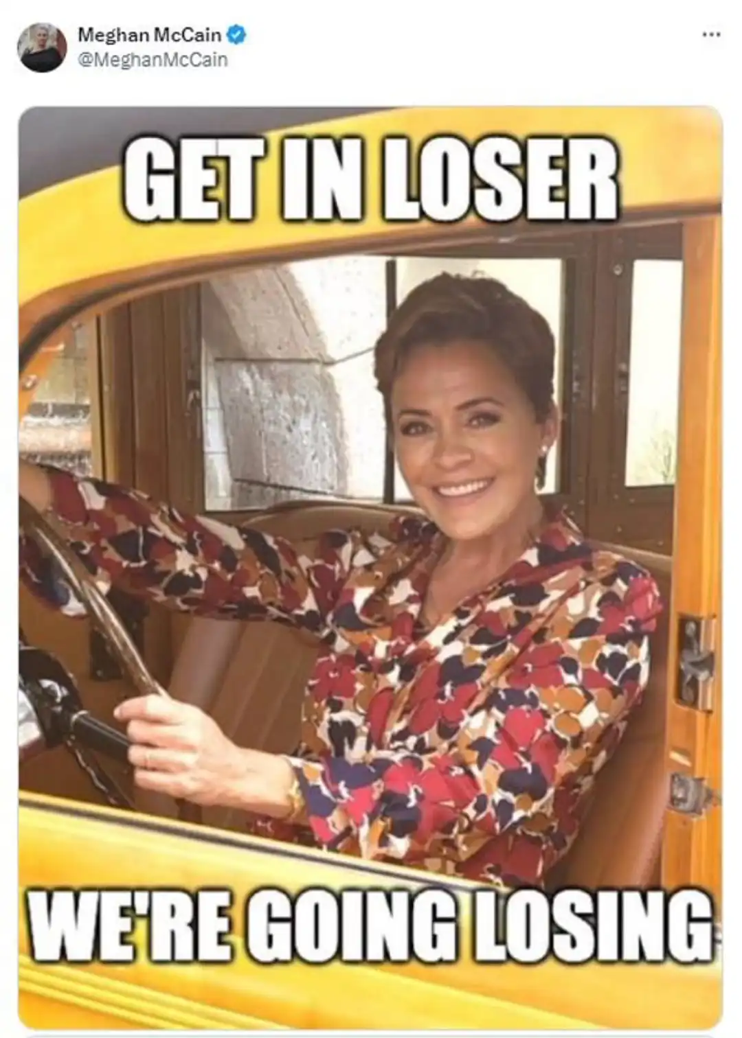 A woman with short brown hair, wearing a colorful patterned blouse, is smiling while driving a yellow vintage car. Large…
