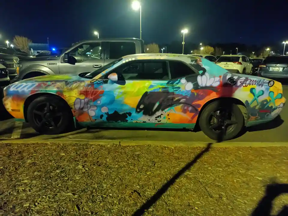 A brightly colored Dodge Challenger covered in vibrant, chaotic graffiti art is parked in a nighttime lot. The car's entire…