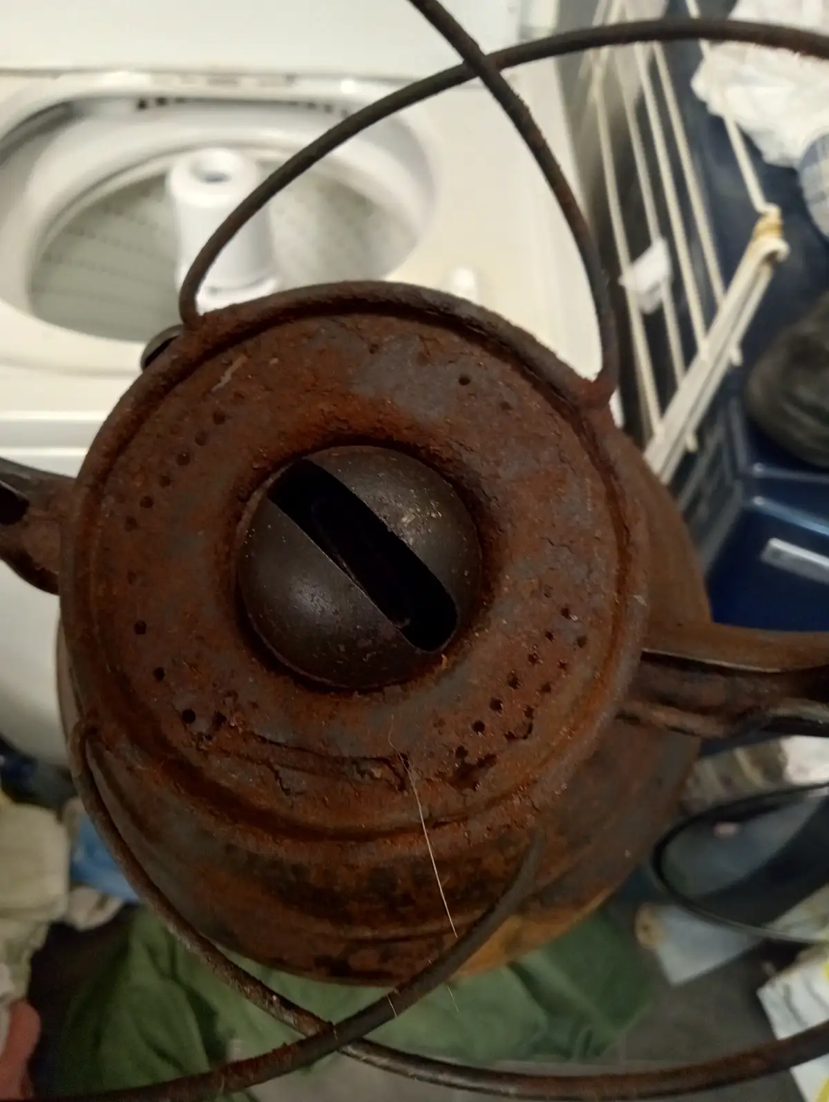 A close-up photo shows a rusty, old metal watering can with a handle and spout, its top viewed from above. The can's lid has…