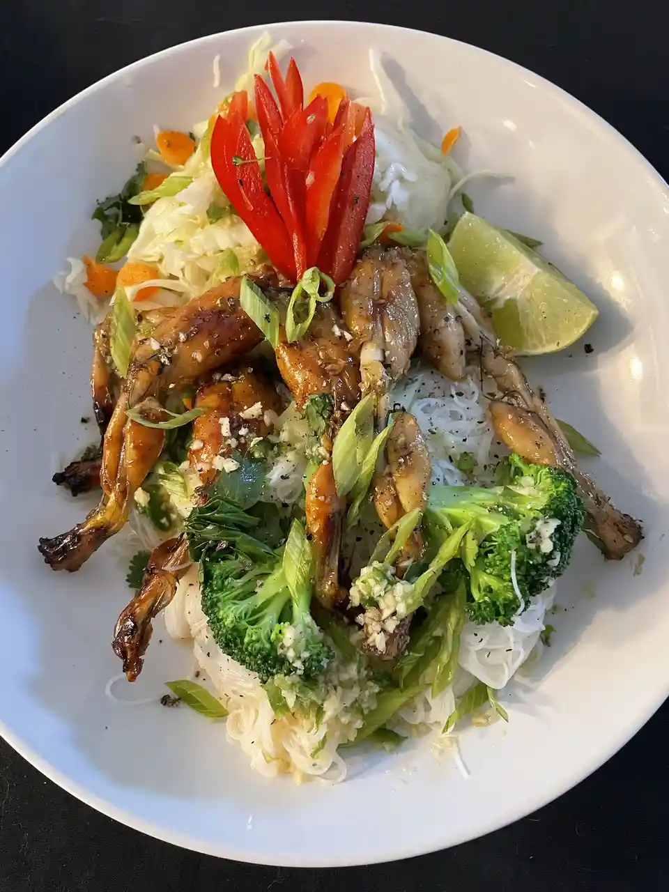 A white bowl contains a dish with cooked frog legs, rice noodles, broccoli, shredded cabbage, carrots, lime wedge, and…
