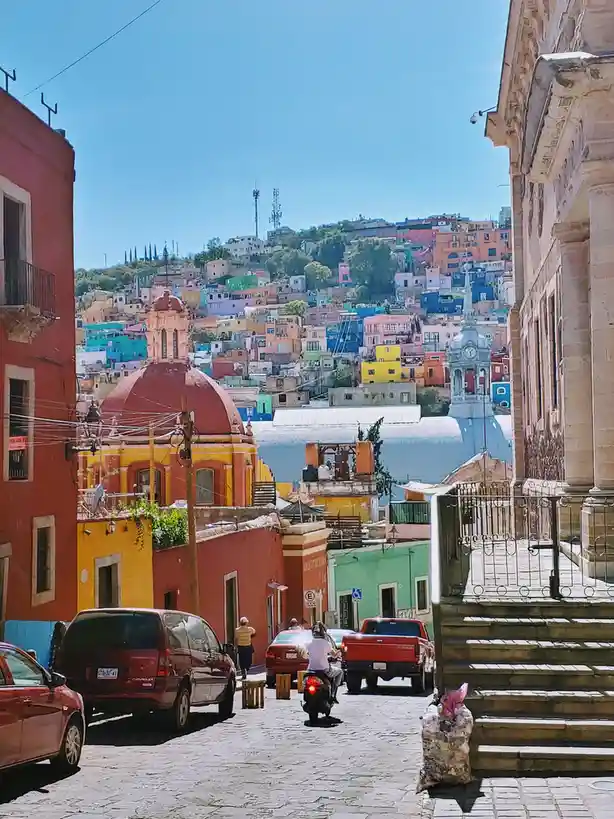A vibrant, sunlit street in a colorful hillside town with multi-hued buildings, a red-domed church, and a scooter rider. The…