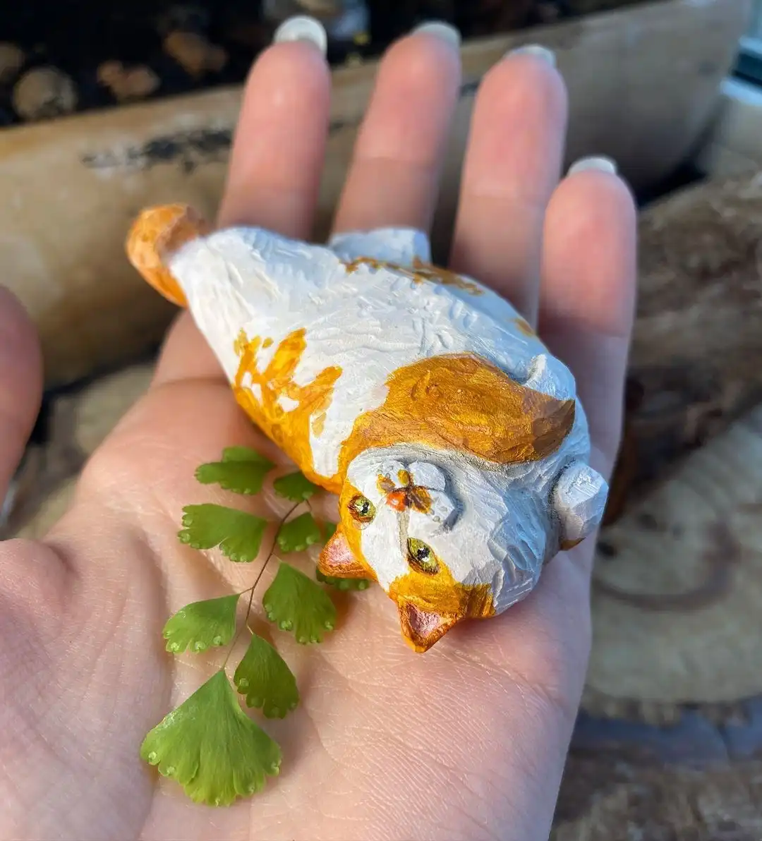 A hand holds a small, hand-painted wooden sculpture of a cat lying on its back, painted white and orange with detailed eyes…