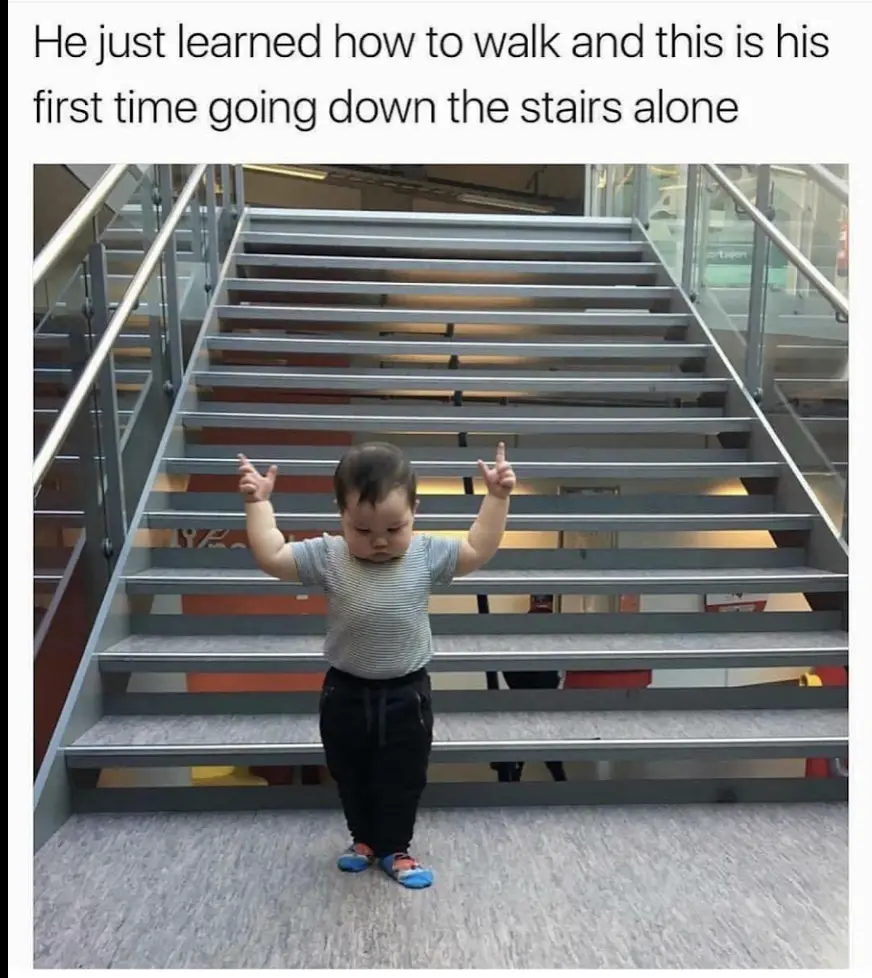 A toddler wearing a striped shirt and dark pants stands at the bottom of a wide, modern staircase with metal railings, arms…