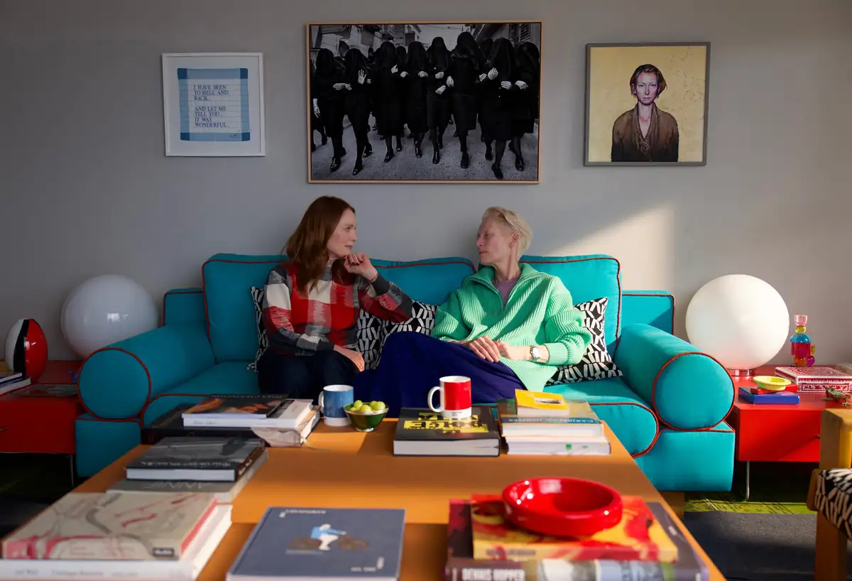 Two women sit on a bright turquoise couch in a colorful, art-filled living room. The wall behind them displays three framed…