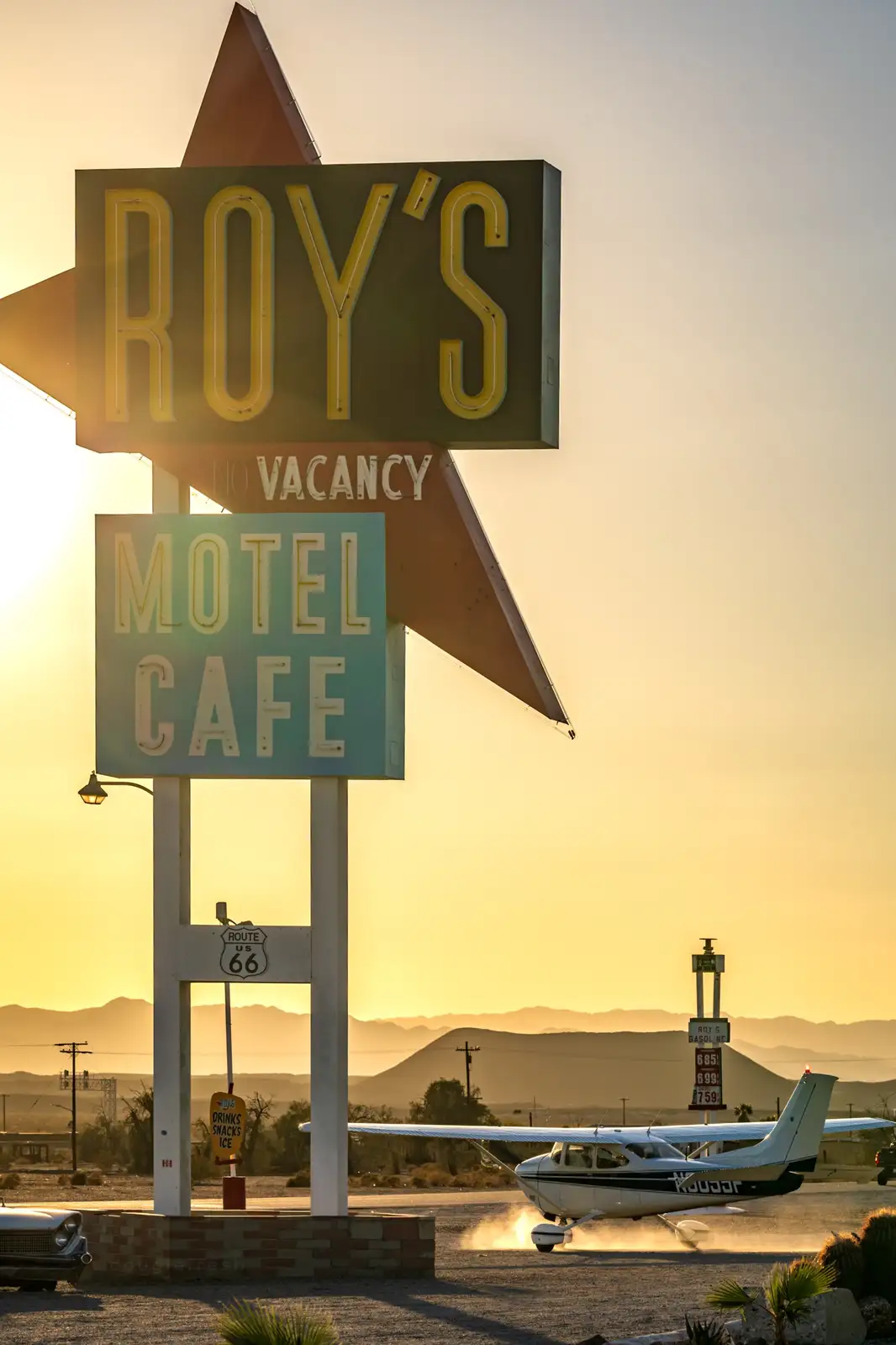 A vintage roadside motel sign for 'Roy's Motel Cafe' with a 'no vacancy' notice, set against a desert sunset with a small…