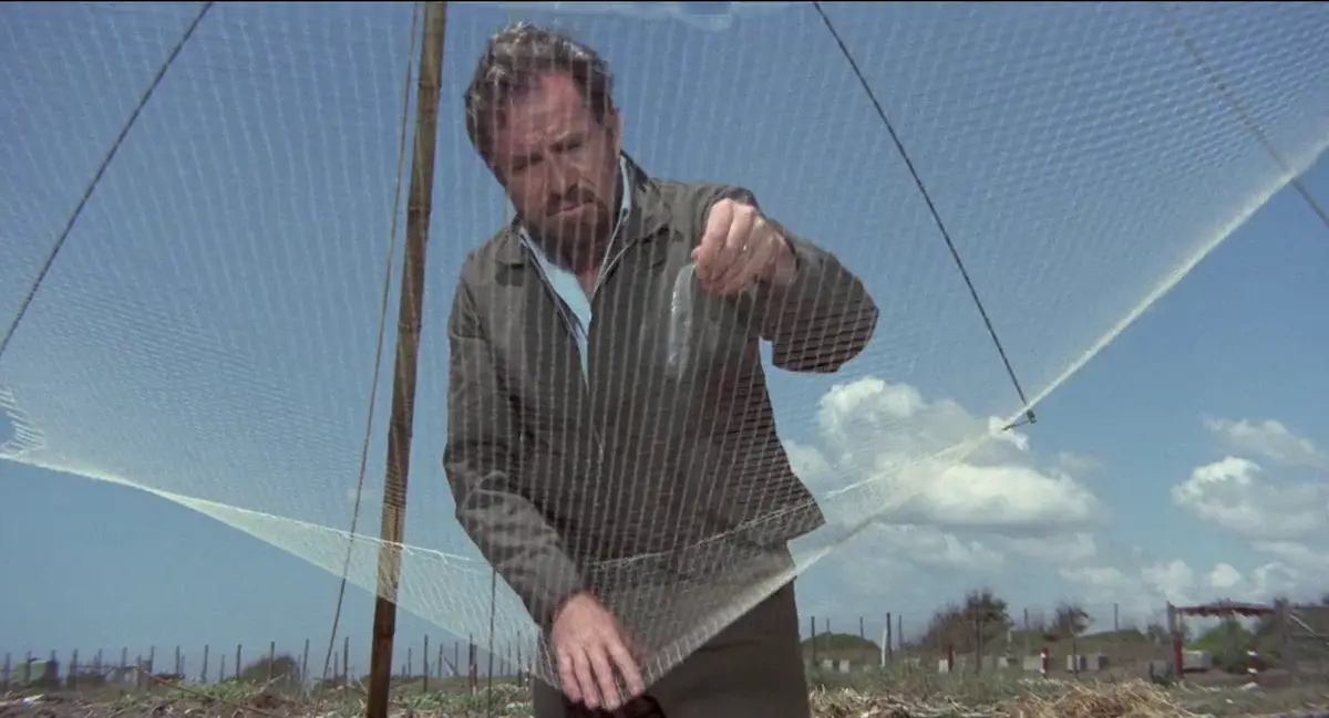 A man in a field, holding a small fish in a transparent plastic bag, stands under a large white net stretched between poles…