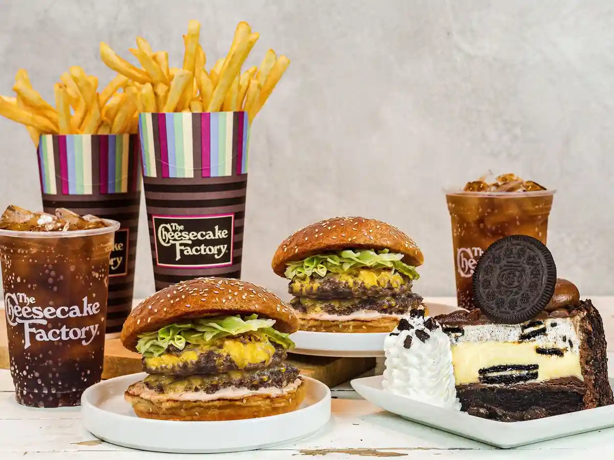 A promotional photo displays a variety of food items from The Cheesecake Factory, including two large cheeseburgers on…