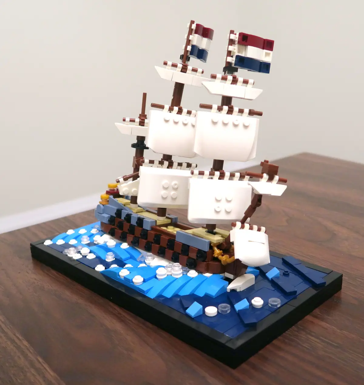 A detailed LEGO model of a multi-masted sailing ship with white sails and red, white, and blue flags, built on a black…