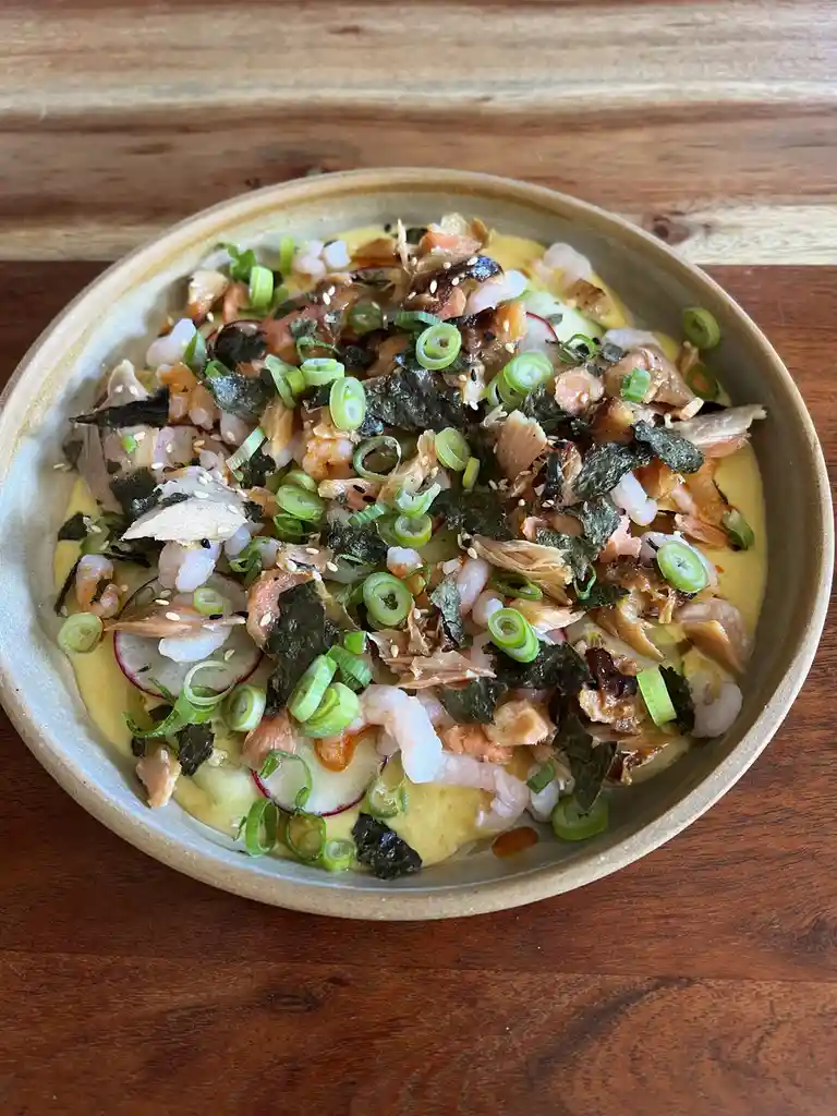 A top-down photo of a ceramic bowl filled with a colorful dish featuring salmon, shrimp, sliced radishes, green onions, nori…
